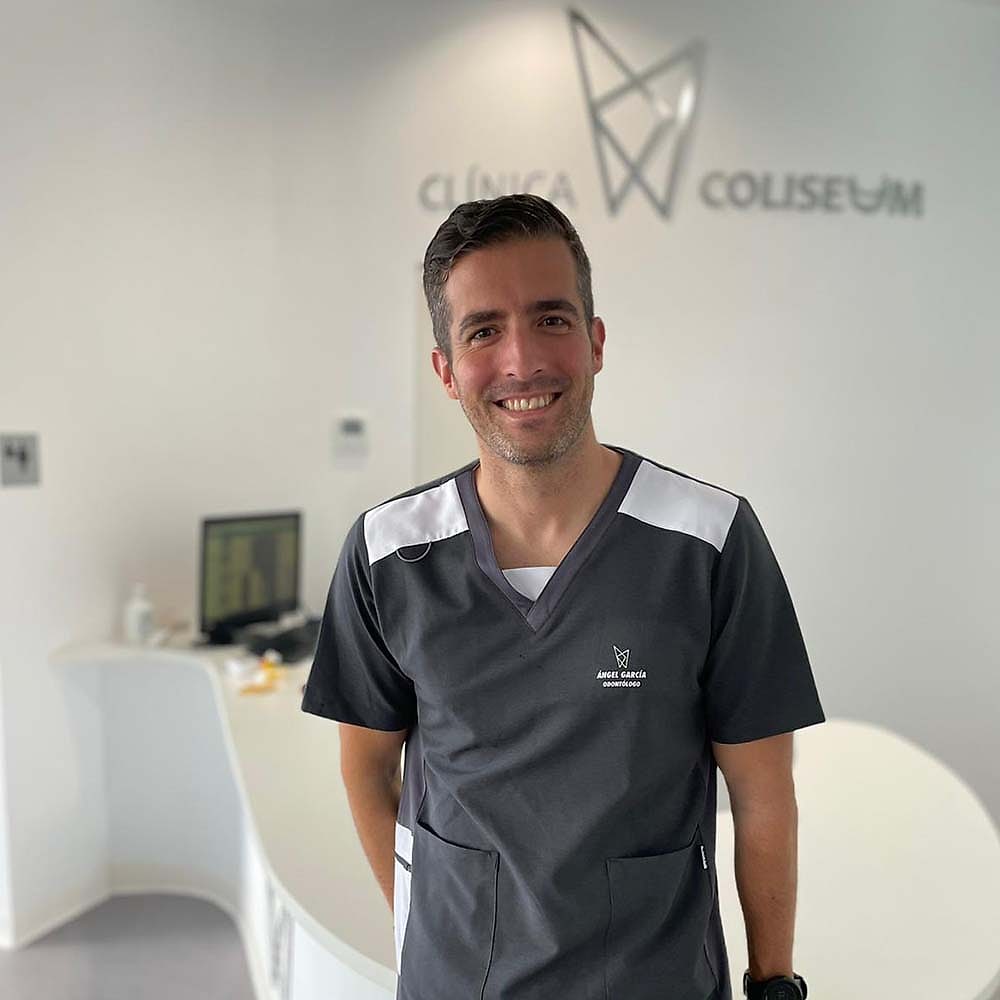 El Dr. Ángel J. García, con uniforme sanitario gris, en la recepción de Clínica Coliseum con el logotipo de la clínica al fondo